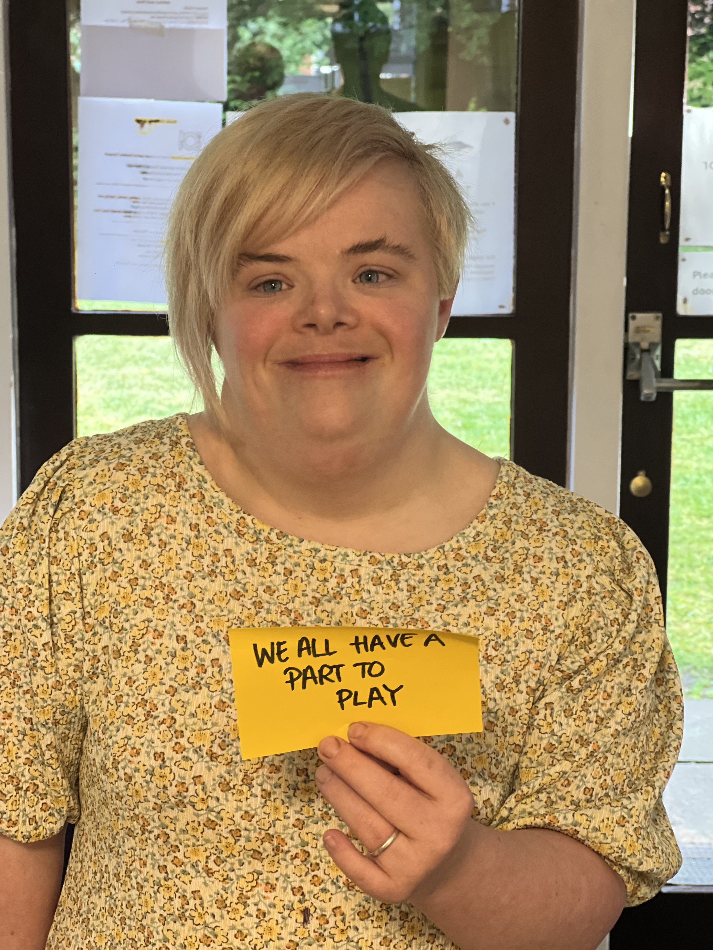 A photo of Heidi holding a sign that says "We all have a part to play" Heidi is wearing a floral dress.