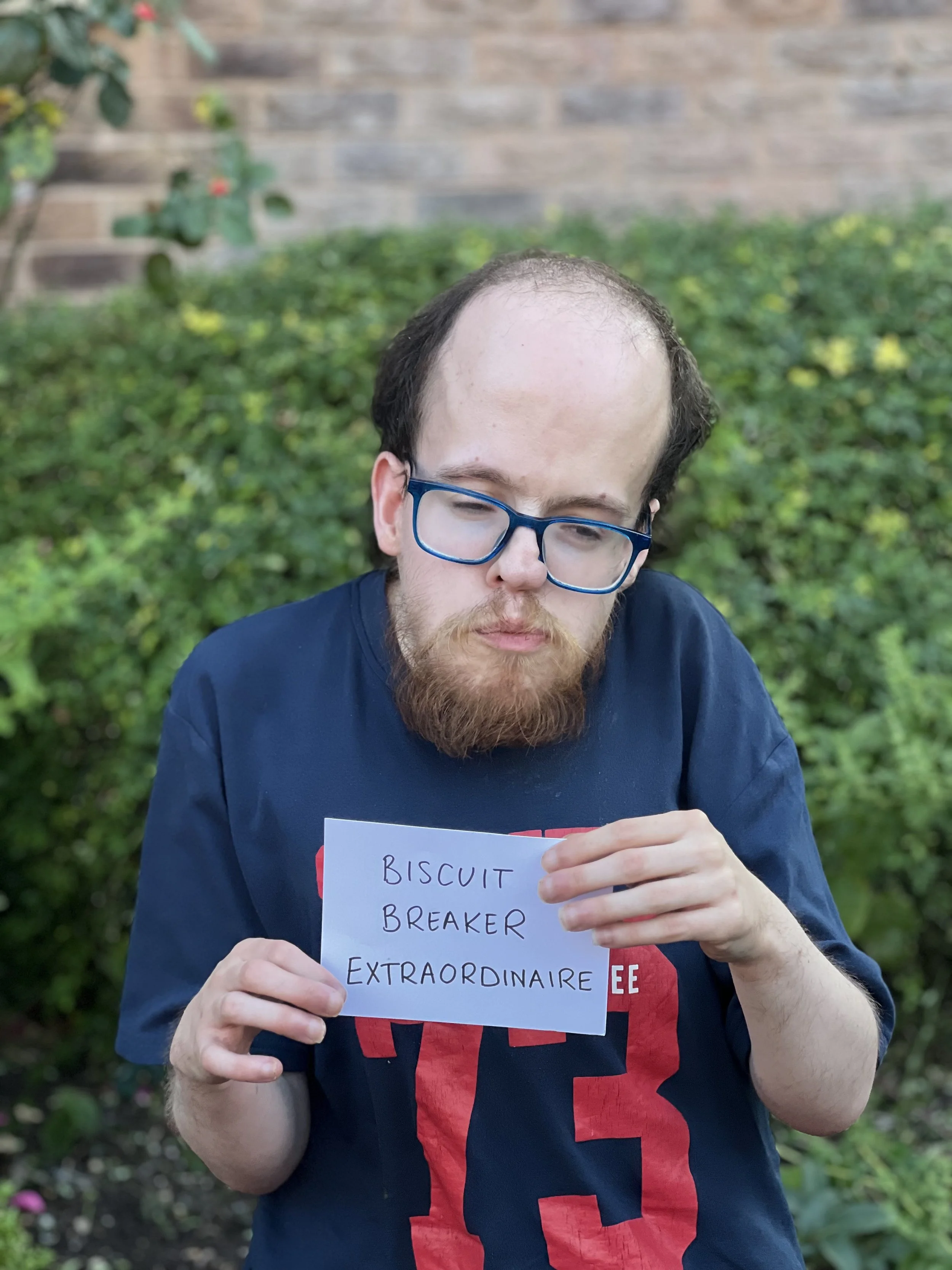 A photo of Tom, wearing blue glasses. Tom is holding a sign that says "Biscuit Breaker Extraordinaire"