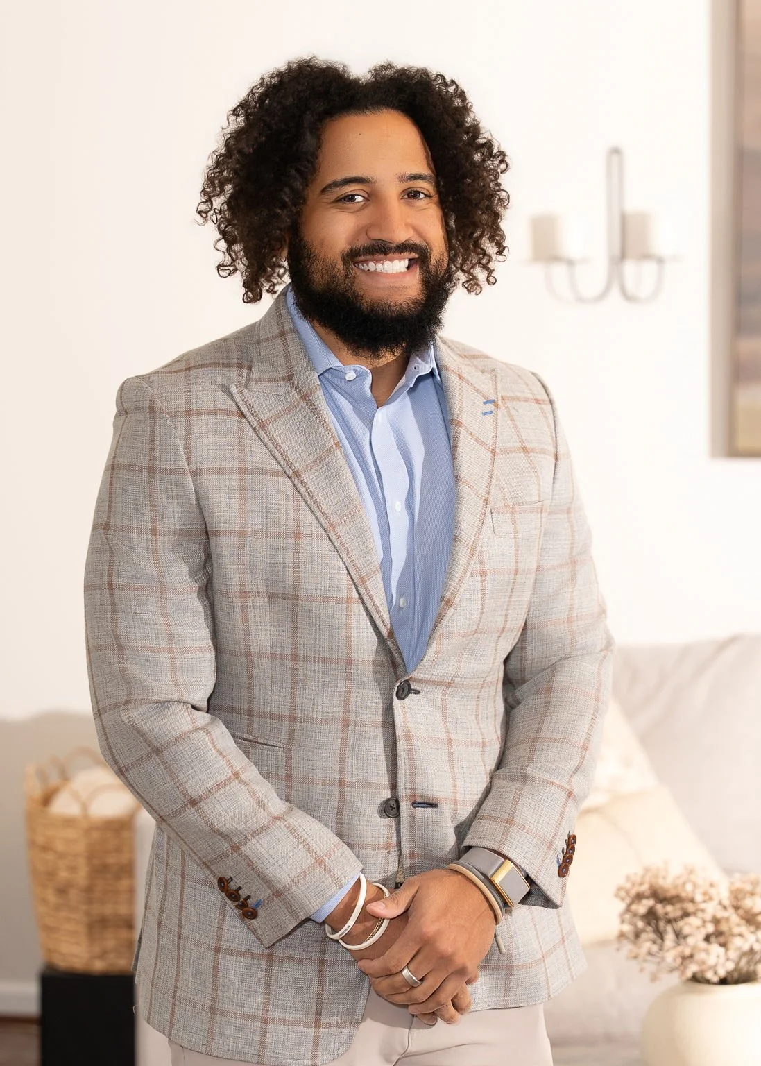 A smiling man with curly hair and a beard, wearing a beige suit and light blue shirt, sitting on a green chair against a dark curtain background.