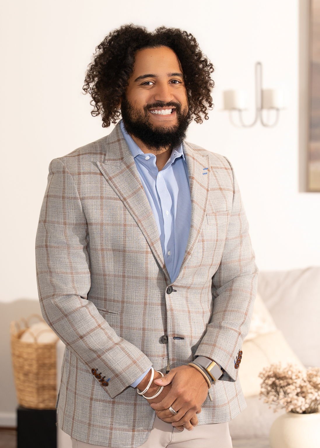 A smiling man with curly hair and a beard, wearing a beige suit and light blue shirt, sitting on a green chair against a dark curtain background.