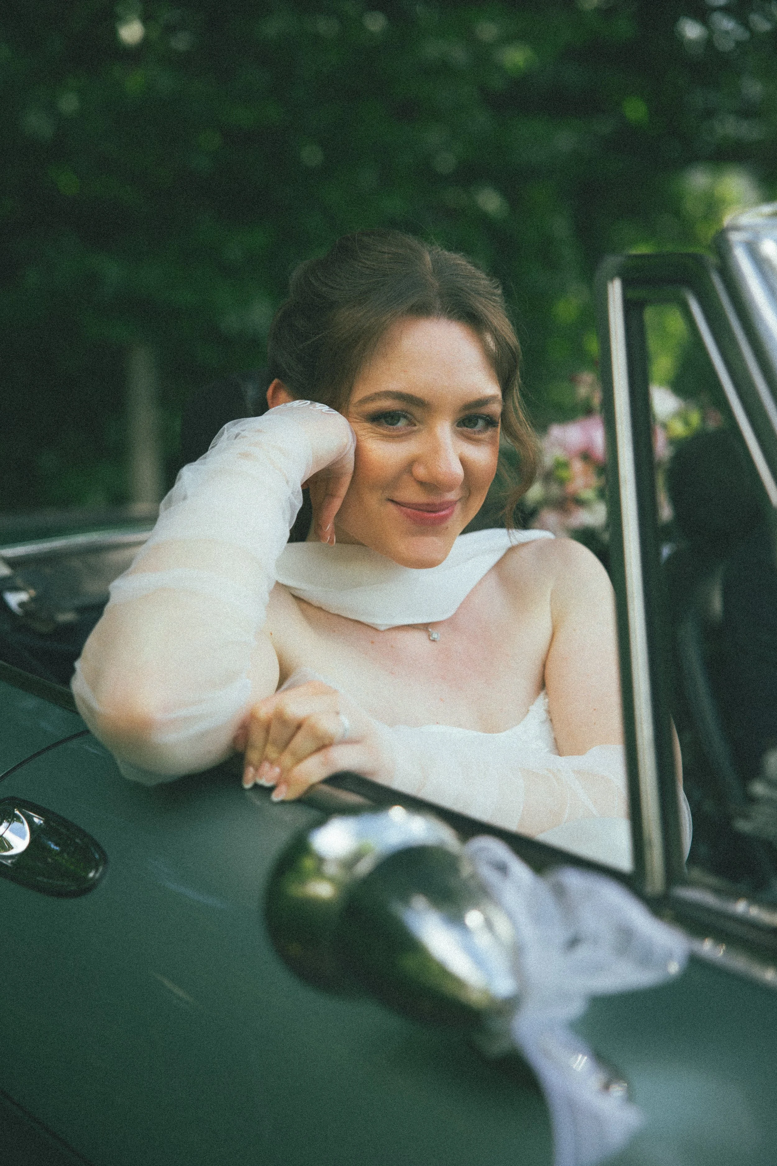 Ein Lächeln einer jungen Frau, die aus einem schwarzen Auto heraus schaut, trägt ein weißes Brautkleid und sitzt im Auto. Es scheint eine Hochzeit zu sein.
