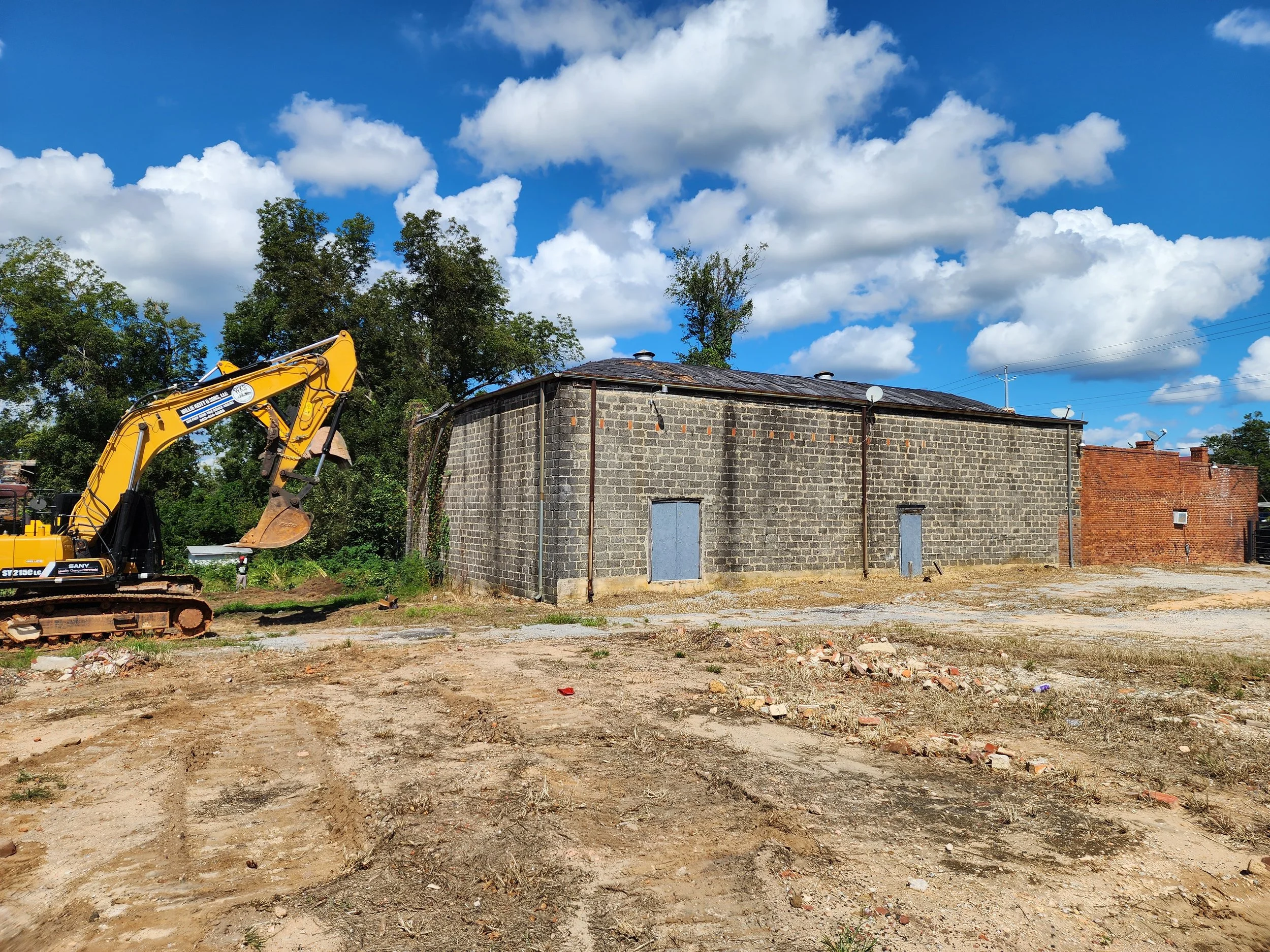 An excavation site with a yellow excavator on the left, a gray brick building with boarded windows in the center, and a red brick building in the background under a partly cloudy blue sky.