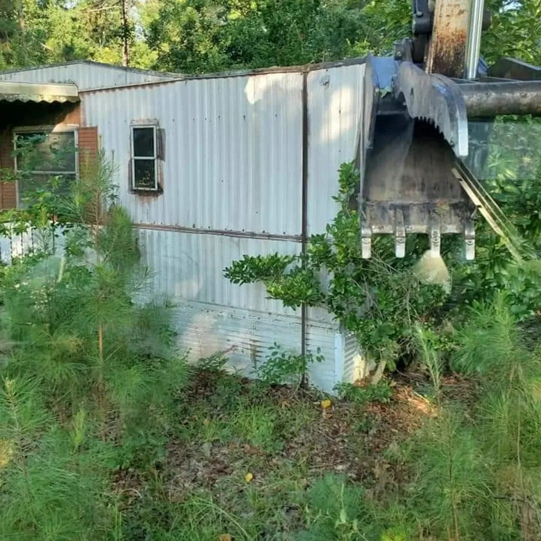 A small metal building with a rusted window and door, surrounded by green trees and bushes, with a large piece of construction equipment or machinery partially visible on the right.