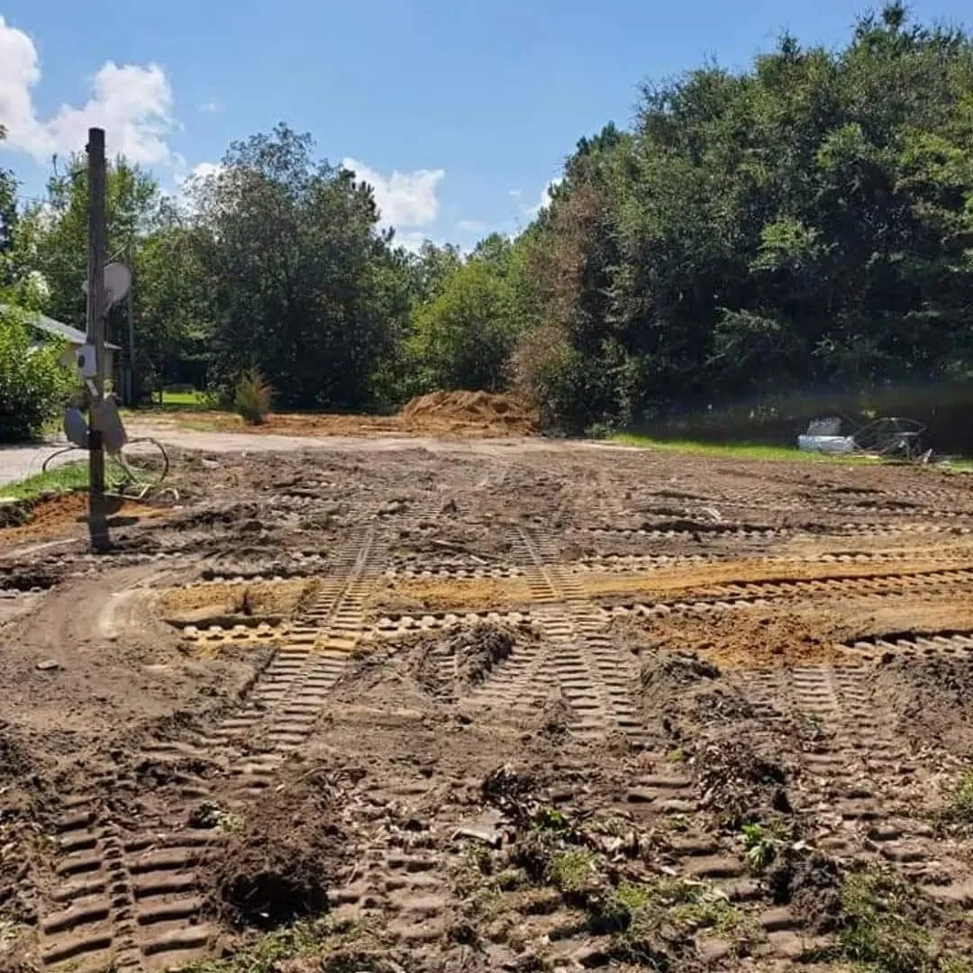 A dirt road under construction with visible tire tracks and graded earth, surrounded by trees and greenery, with a partly cloudy sky overhead.