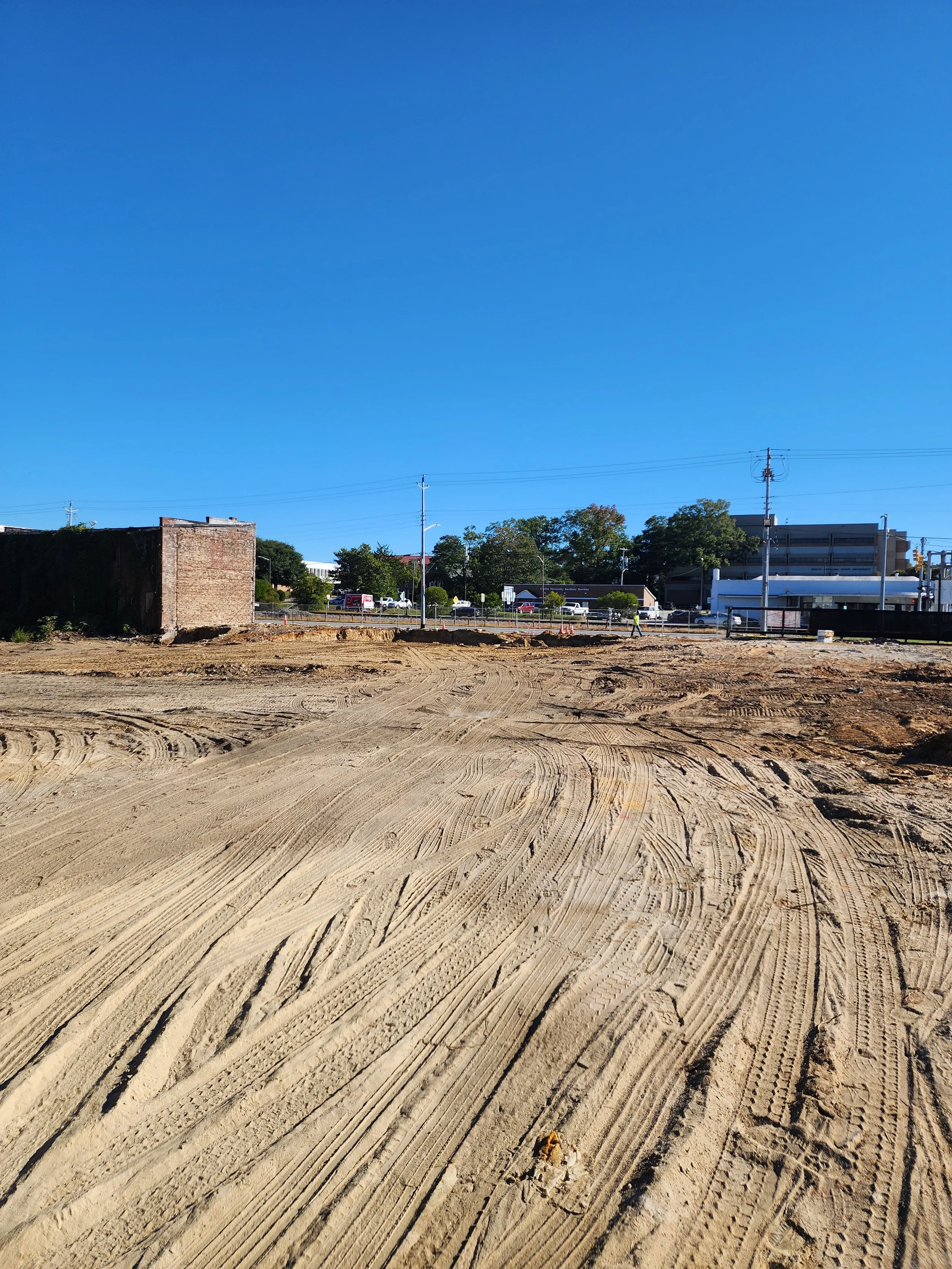 Empty construction site with tire tracks in the dirt, and buildings, trees, and power lines in the background under a clear blue sky.
