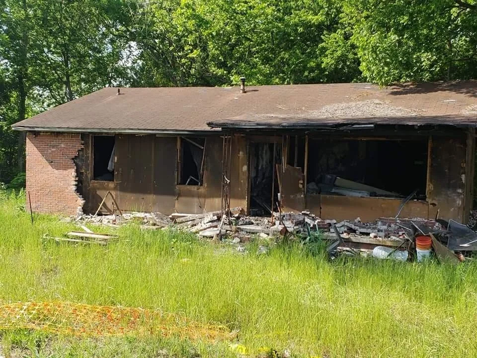 A house with extensive fire damage, with charred walls and broken windows, surrounded by green grass and trees.