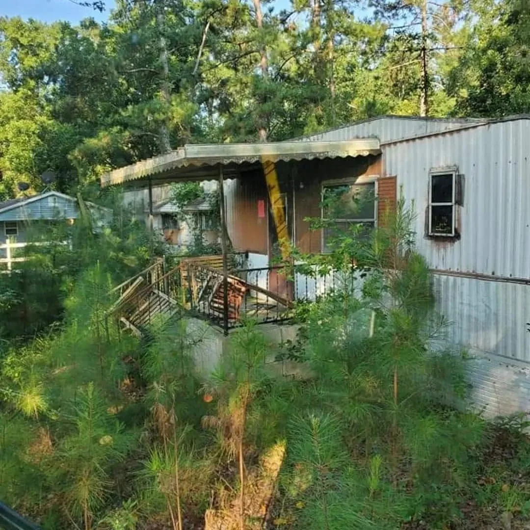A small house or mobile home with a metal exterior, surrounded by trees and greenery, with a small wooden deck at the front. The house shows signs of fire damage, with blackened windows and a burnt wall.