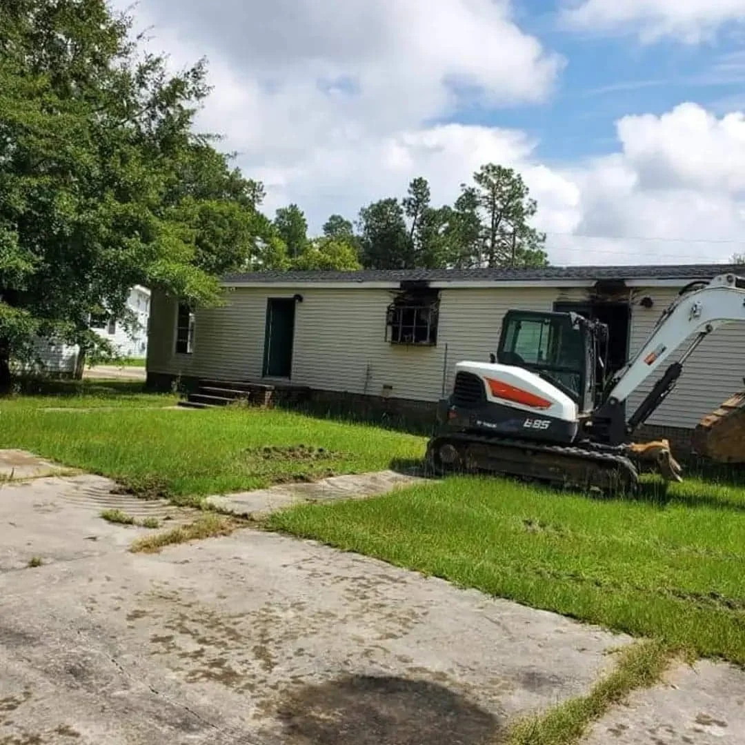 A house with blackened windows and part of the wall burned, surrounded by green grass and trees, with a small excavator parked nearby.
