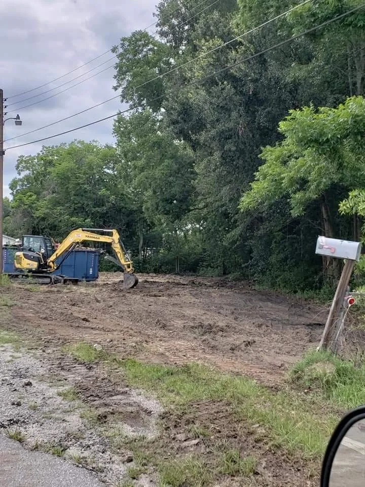 A yellow excavator clearing a dirt driveway next to a green tree and utility pole with overhead power lines, in a semi-rural area.