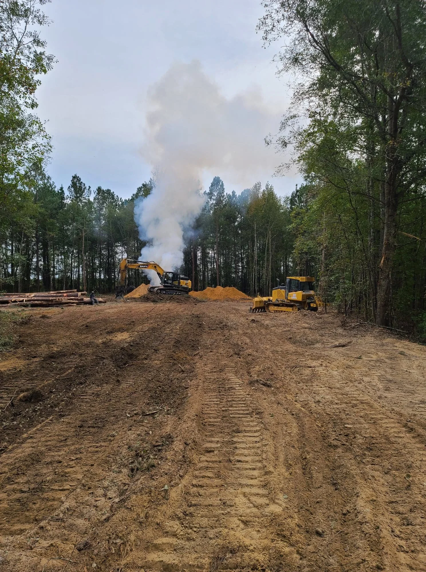 Construction site with two yellow bulldozers and a backhoe, dirt ground, trees in the background, and smoke rising from one of the machines.