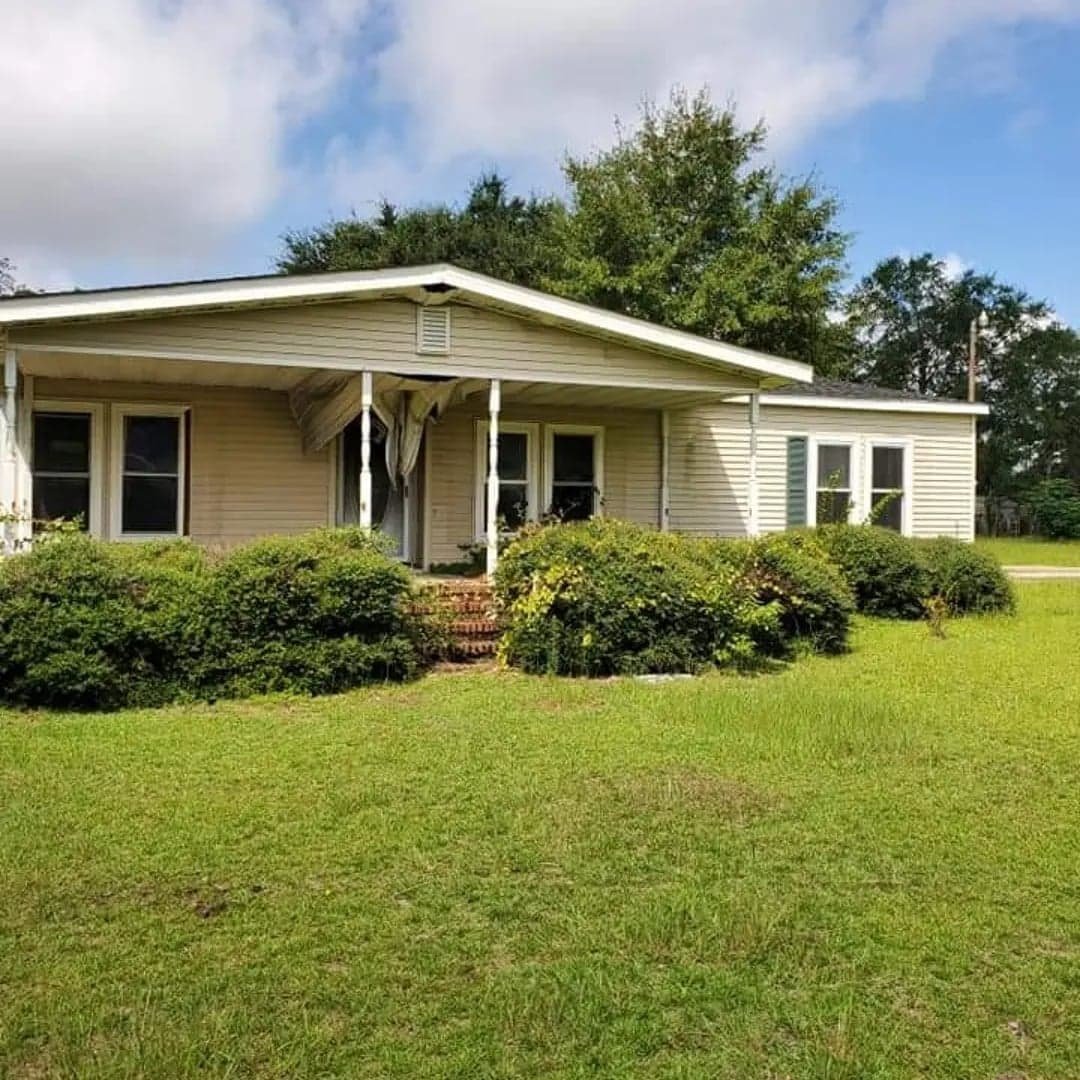 A single-story house with beige siding, a small front porch with white supports, and trees in the background. The yard has green grass and bushes.