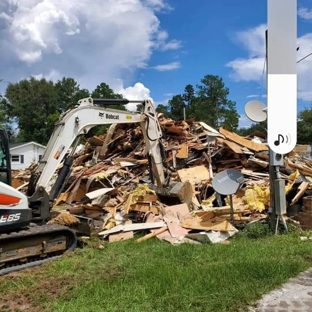 A pile of debris from a demolished building in a yard with a small Bobcat excavator working, and communication satellite dishes in the scene on a sunny day