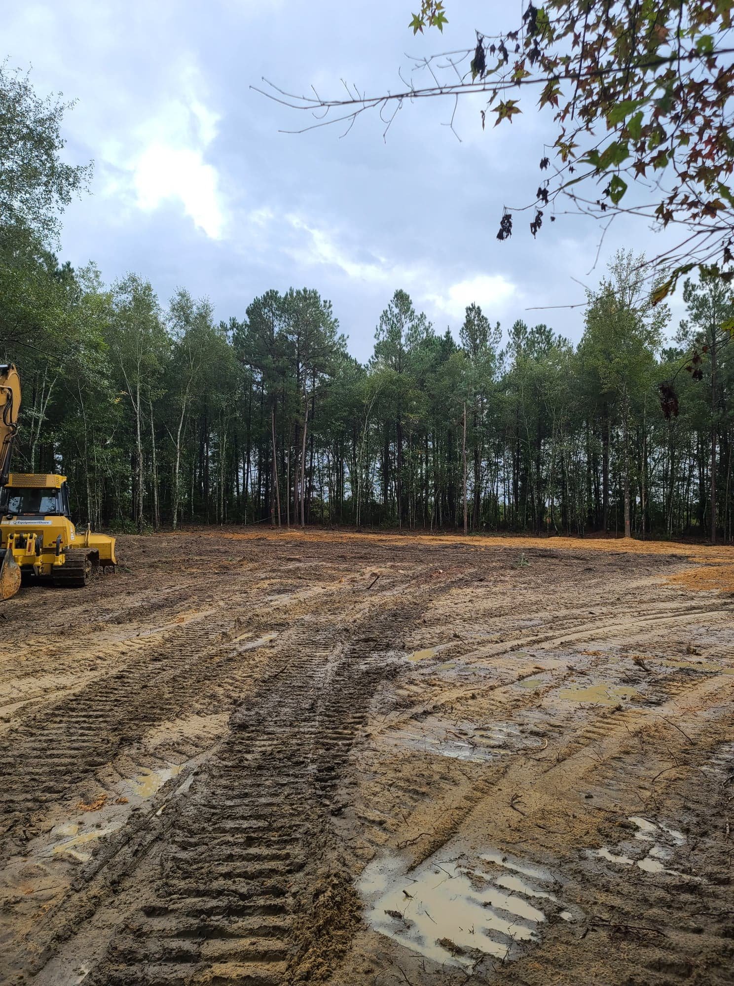 A cleared construction site with muddy tire tracks, some puddles, and a yellow bulldozer on the left, with a background of green trees and a cloudy sky.