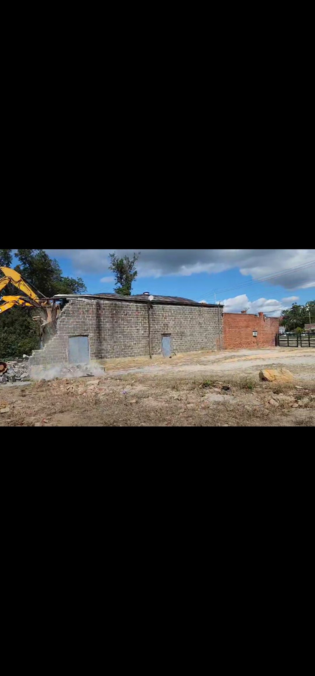 A house under demolition with a yellow excavator tearing down a brick wall, dirt and rubble on the ground, and a partly cloudy sky overhead.