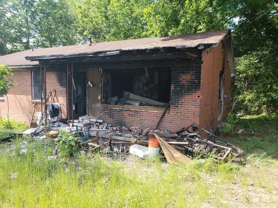 A house with extensive fire damage, including a charred and partially collapsed front porch and burnt interior, surrounded by green grass and trees.