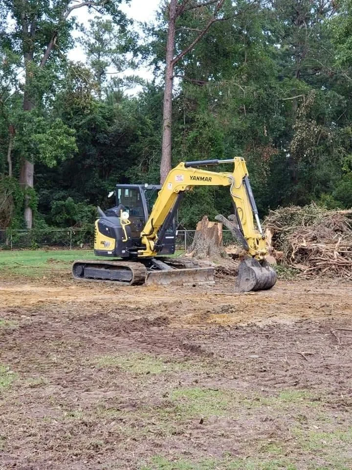 A yellow and black excavator working on clearing wood debris in a yard with trees and a fence in the background.