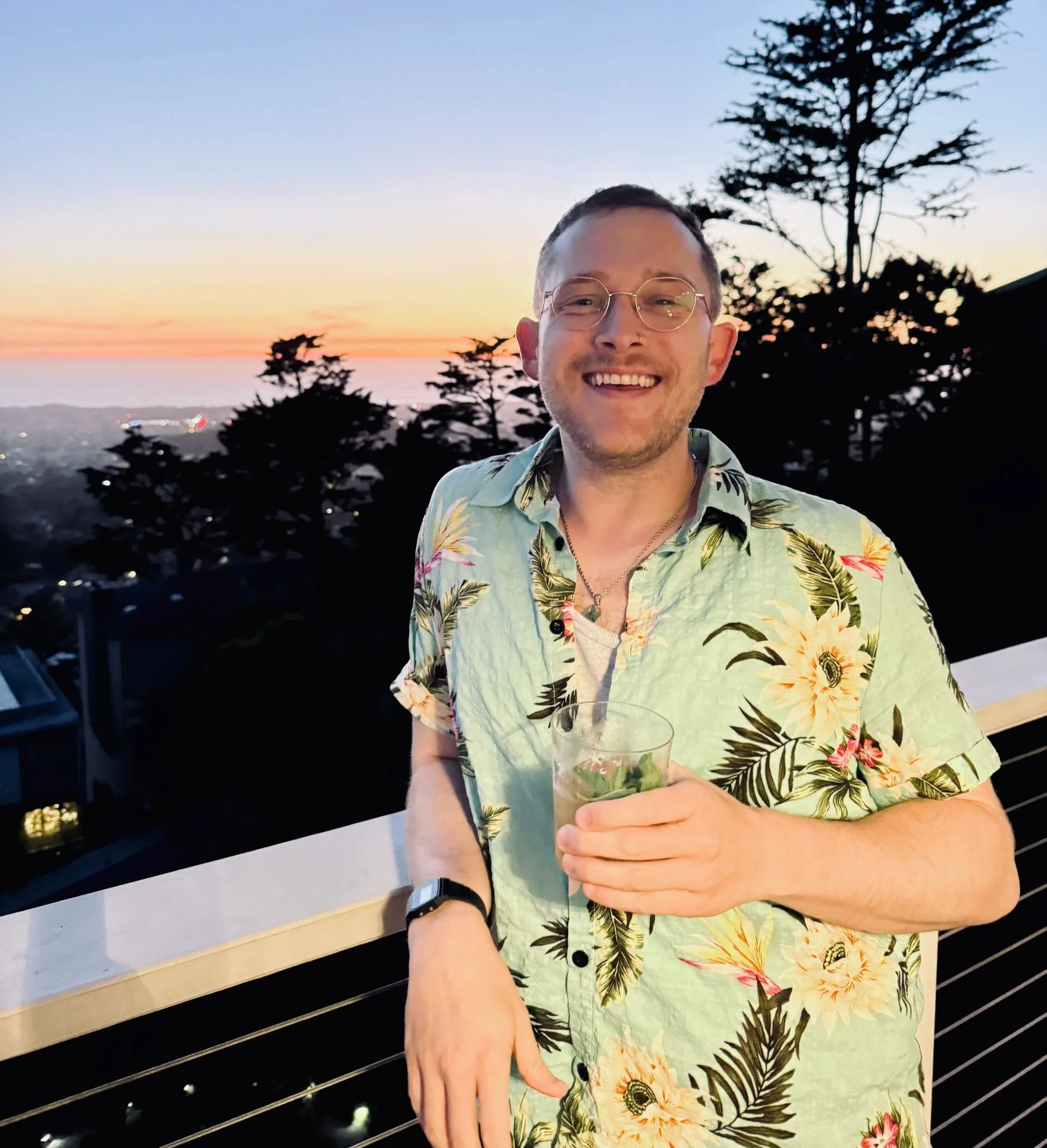 Person with short dark hair and glasses smiling, wearing a Hawaiian shirt, holding a glass of a green drink with ice and mint, standing outdoors at sunset with silhouette trees and city lights in the background.