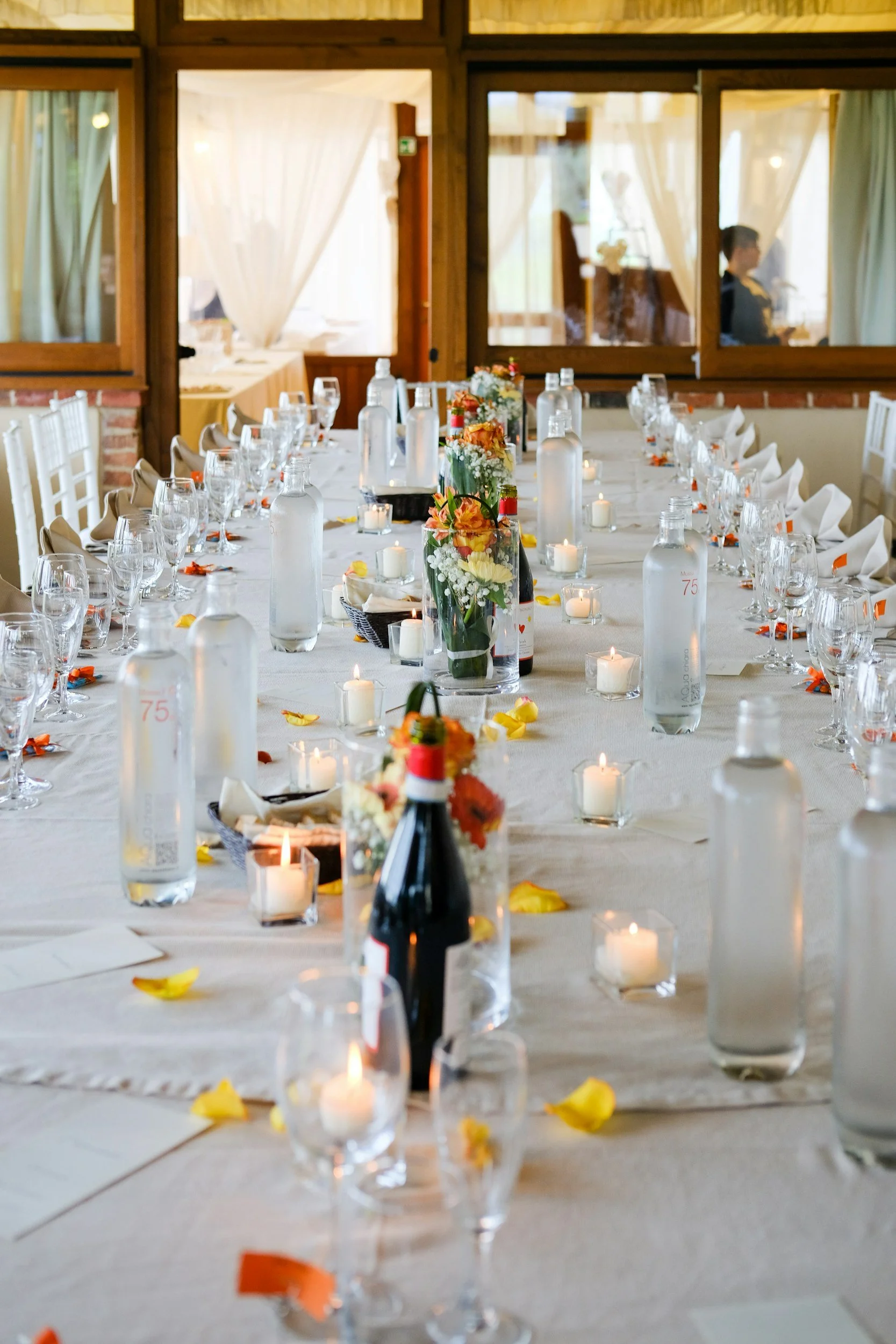 A long banquet table set for an event with white tablecloths, floral centerpieces, wine bottles, water bottles, glasses, candles, and scattered flower petals inside a decorated event space.