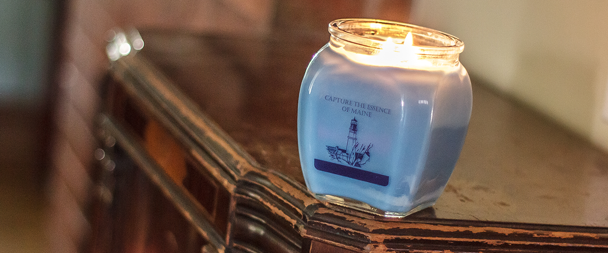 A lit candle in a blue jar with a lighthouse illustration and the text 'Capture the essence of Maine' on a wooden surface.