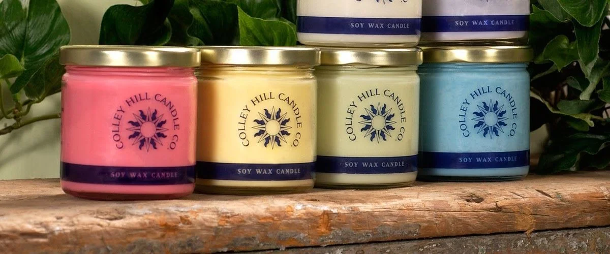 A row of four colored soy wax candles in glass jars with metal lids, placed on a wooden surface with green leaves in the background. The candles are pink, yellow, pale green, and blue, each labeled with 'Colley Hill Candle Co.' and 'Soy Wax Candle'.