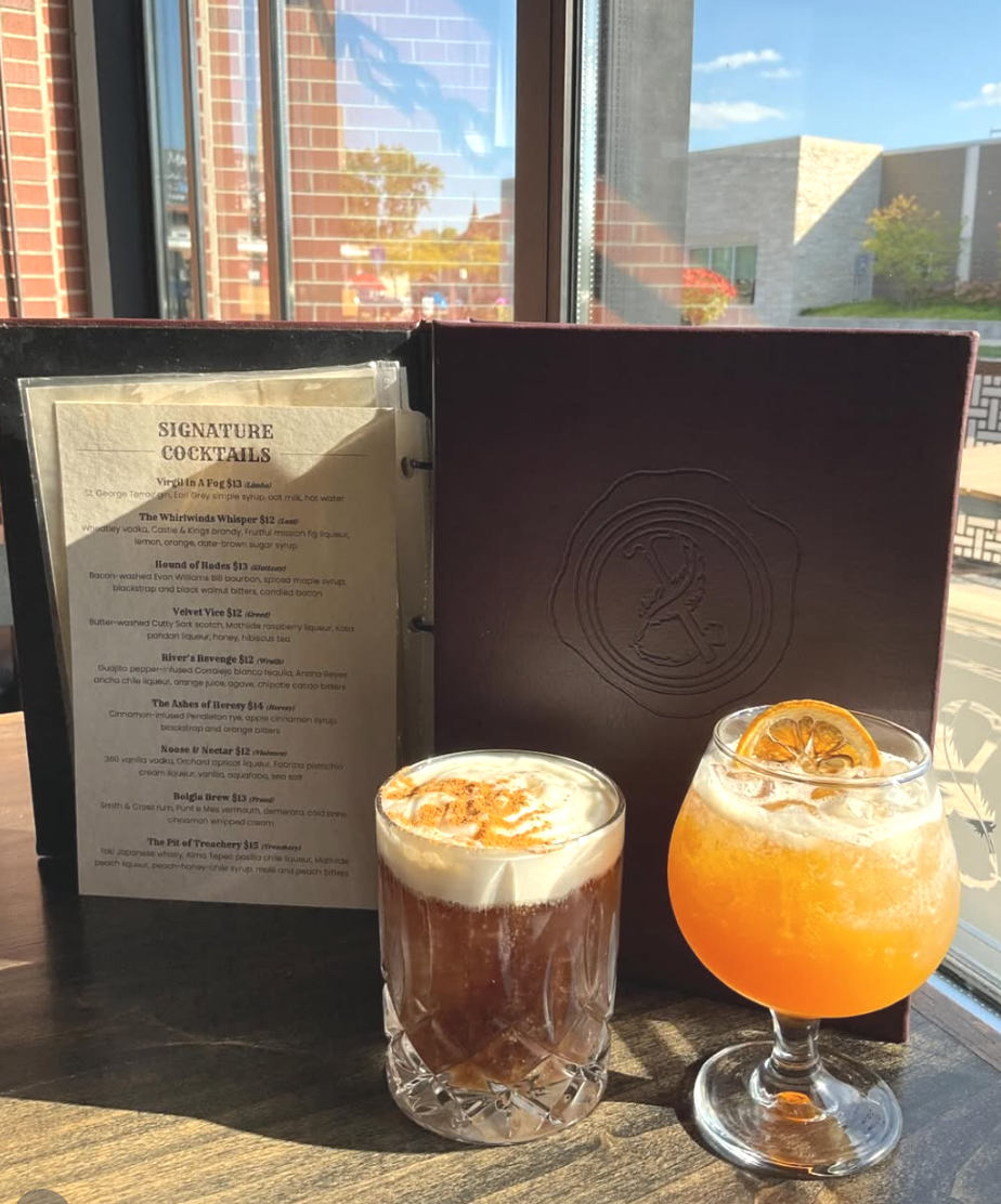 Two cocktails on a wooden table near a window, with a menu in the background. One cocktail is dark with ice and a lemon twist, the other is bright orange with a slice of orange.