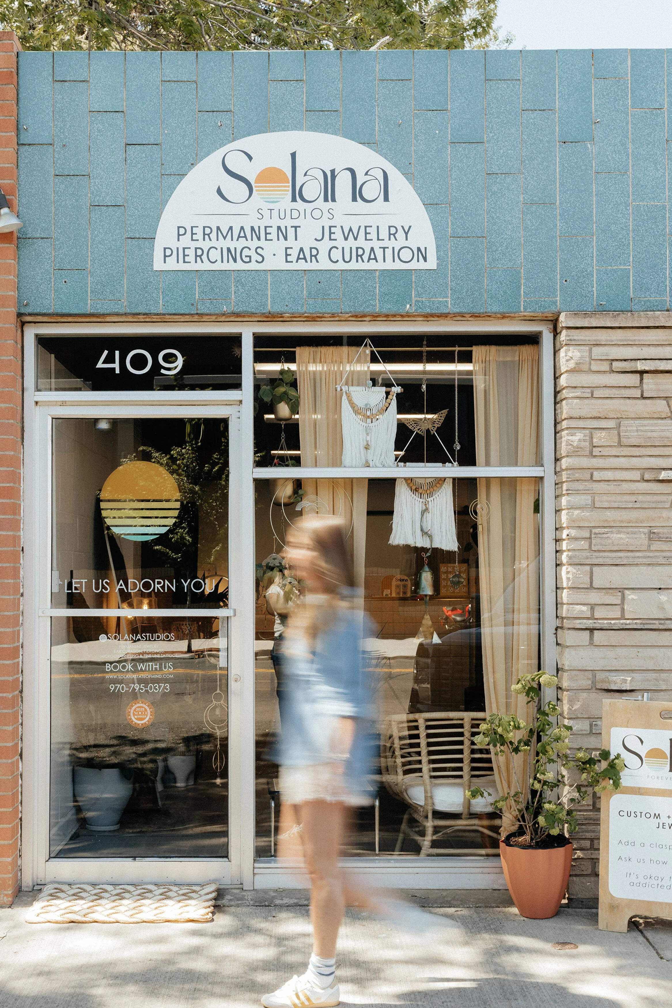 Outside storefront of Solana Studios with signage advertising permanent jewelry, piercings, and ear curation. Large window display shows hanging decor and potted plants inside, with a person walking past in the foreground.