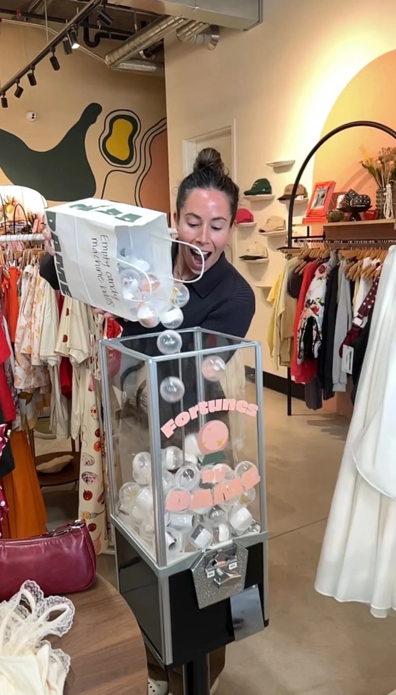 Woman wearing black shirt in a clothing store, playing a game where she drops small plastic balls into a transparent box labeled 'Förtung' and 'Drop', with clothing racks and shelves with hats in the background.