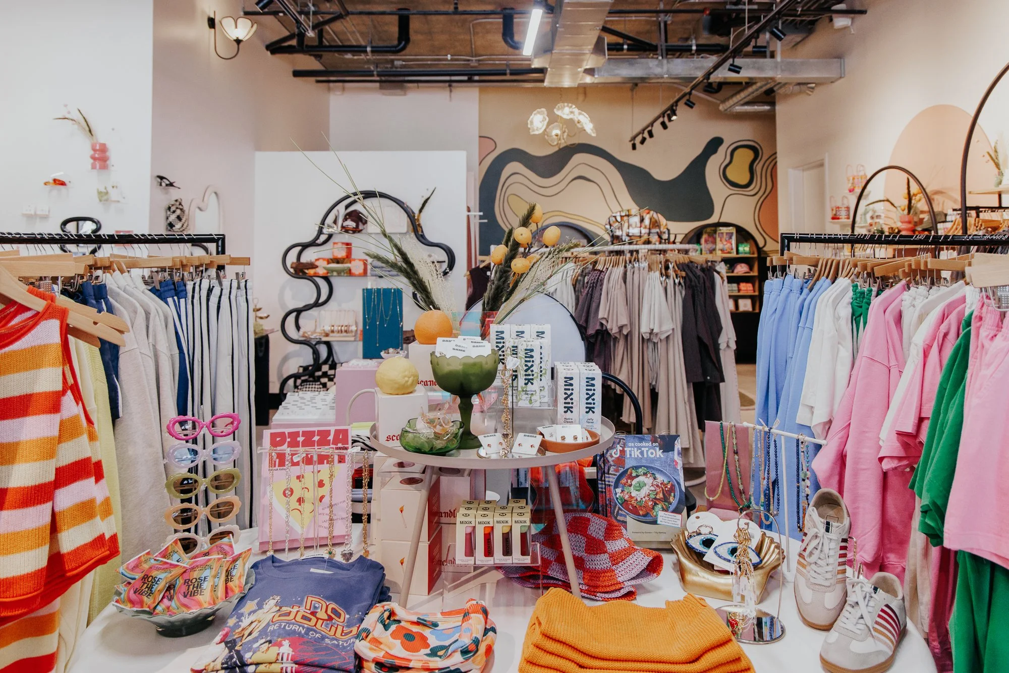 Clothing store display with colorful shirts, accessories, and home decor items on tables and racks, with abstract wall art in the background.