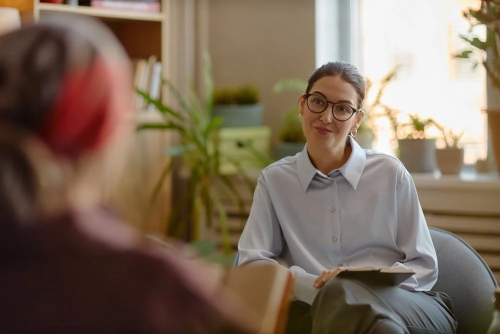 A young woman with glasses and a white shirt sitting in a chair during a conversation in a cozy, well-lit room with plants and a bookshelf.