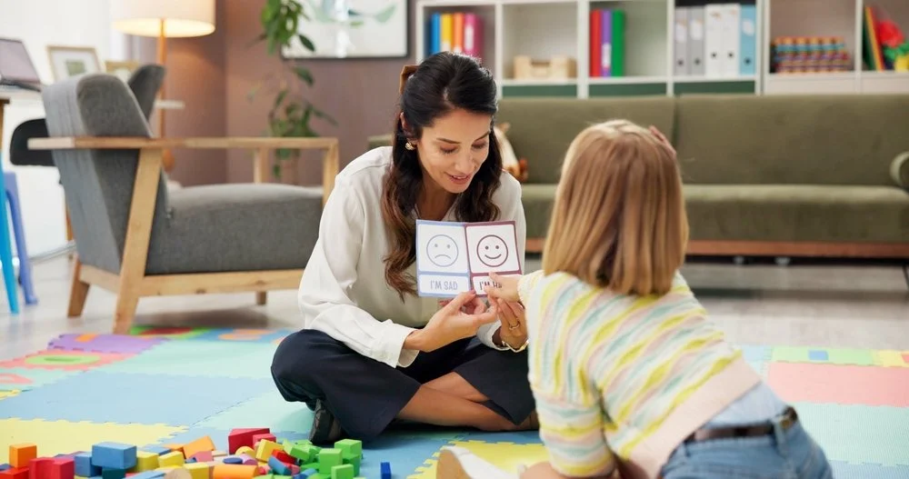 A woman smiling and showing a wooden card with happy face and sad face images to a young girl lying on a colorful foam play mat with building blocks around them, in a cozy living room.