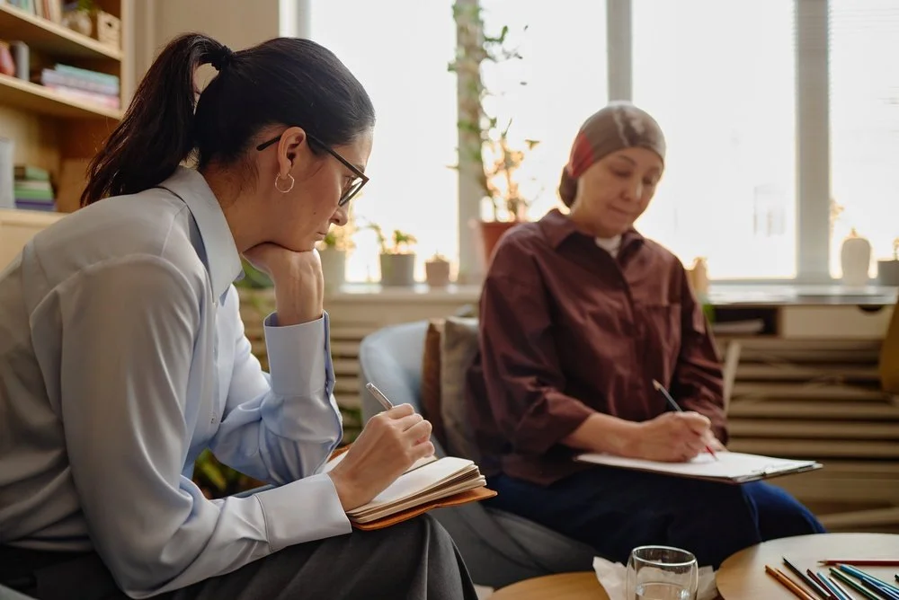 Two women sit indoors, taking notes. One has dark hair in a ponytail, glasses, and a white shirt; the other is wearing a headscarf and a brown blouse. They are in a bright room with bookshelves and windows.