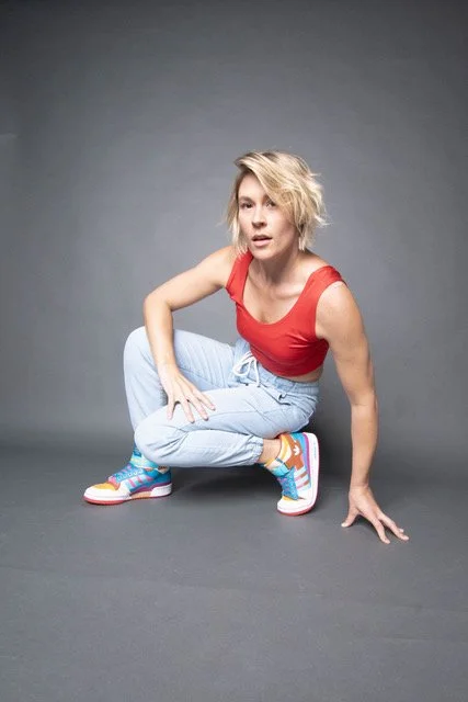 Woman with blonde hair in a red top and light blue jeans at a crouching pose against a gray background.