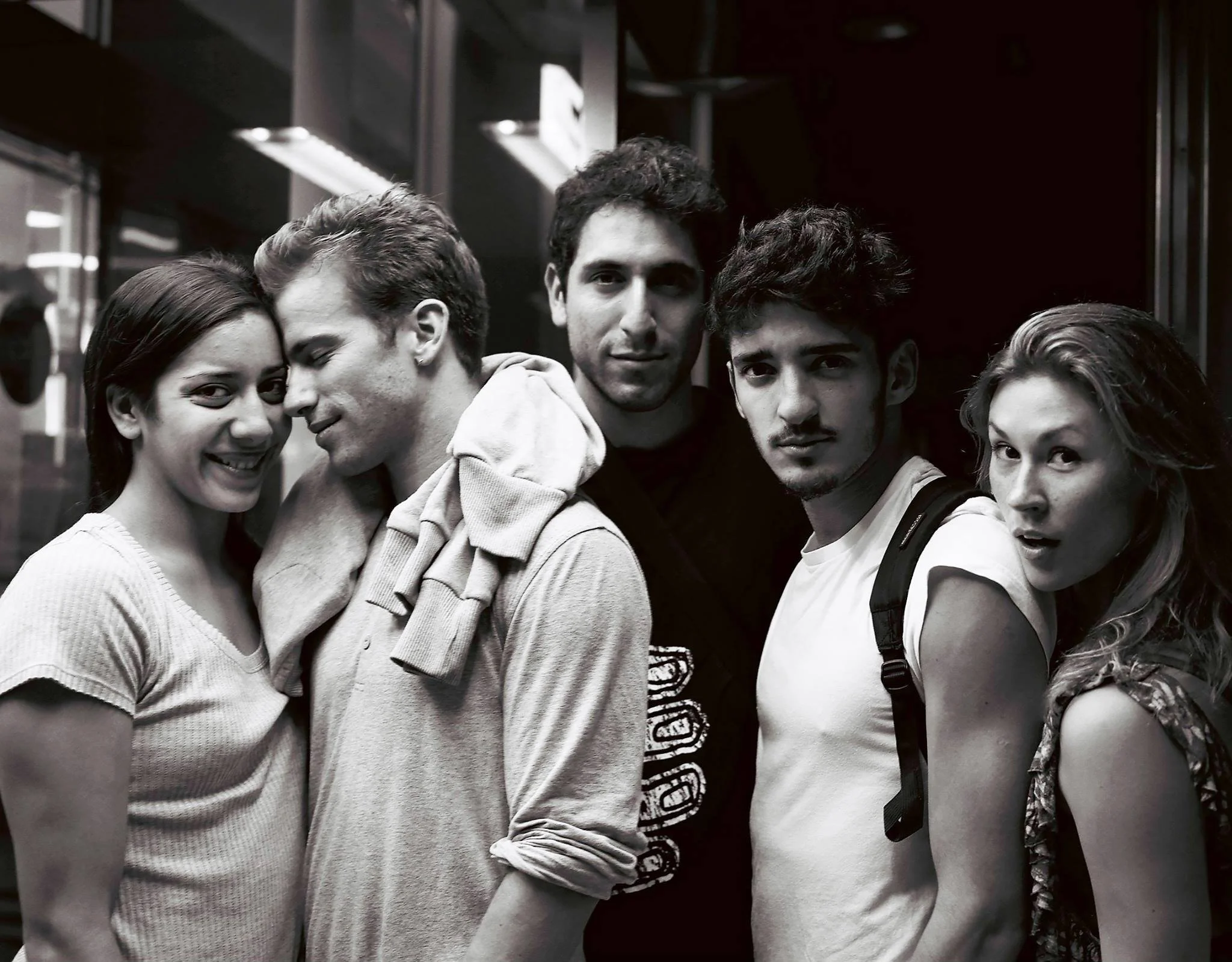 A group of five young people, three men and two women, standing closely together in an indoor setting, with some smiling and others making surprised expressions.