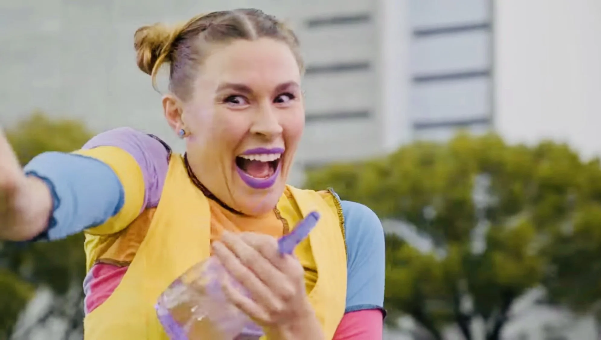 A woman with colorful surroundings, smiling and pointing at the camera, dressed in bright, multicolored clothing and makeup, outdoors with a building and trees in the background.