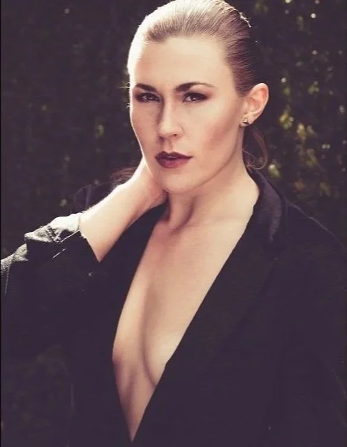 A woman with blonde hair slicked back, wearing dark lipstick, and a black blazer with no shirt underneath, standing outdoors with trees in the background.