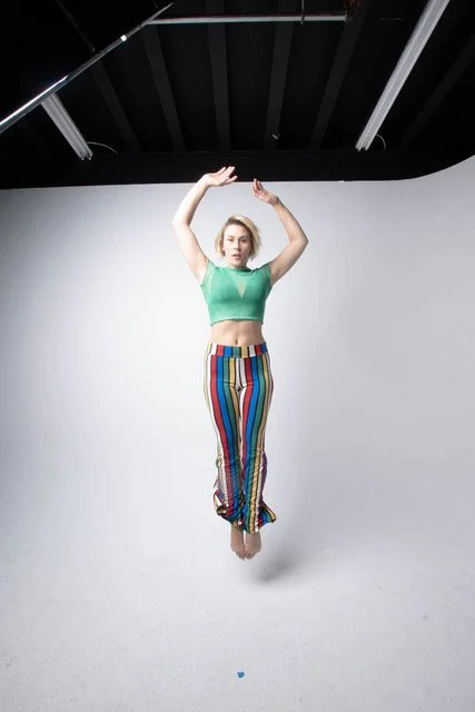 A woman in a green crop top and colorful striped pants appears to be floating or jumping in a studio with a plain white background and a black ceiling.