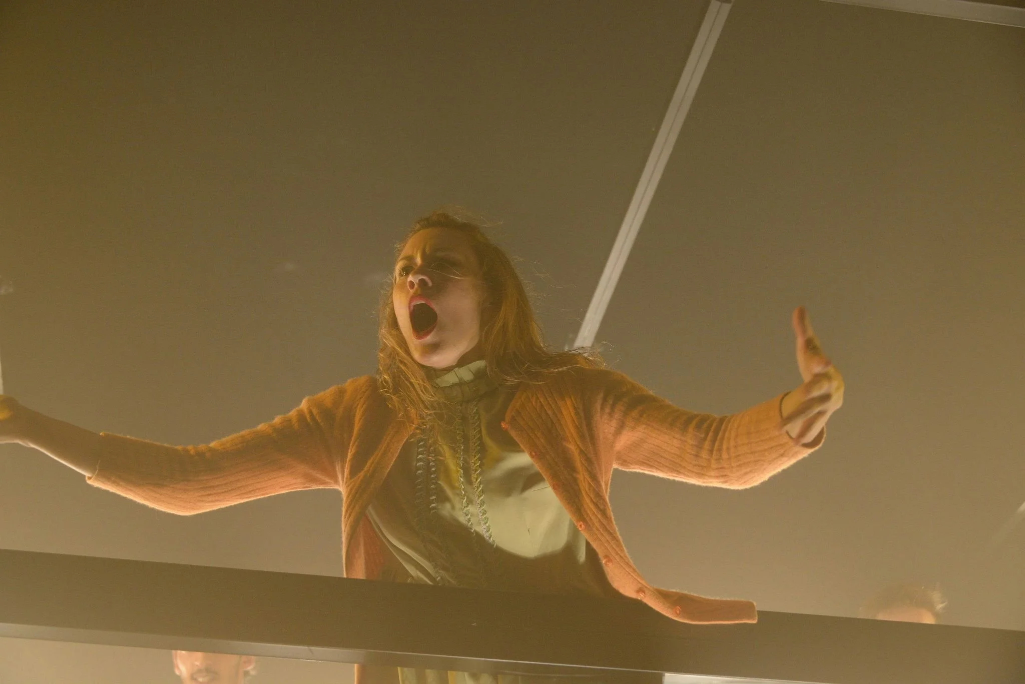A woman with long hair and glasses, wearing a brown jacket, with her mouth open and arms outstretched, looking upward in a dramatic pose inside a dimly lit space.