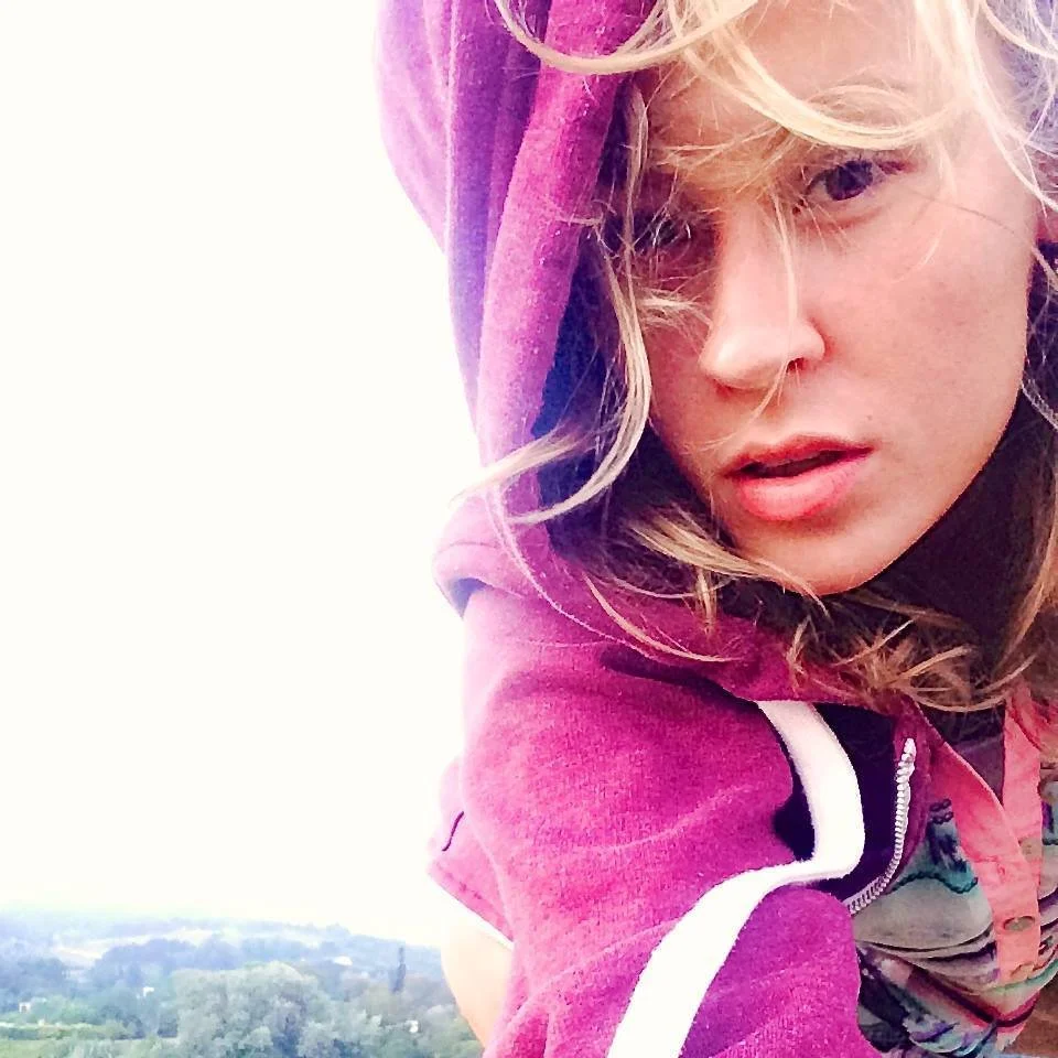 Close-up selfie of a woman with blonde curly hair wearing a pink hoodie, partially covering her face, with a landscape of trees and fields in the background.