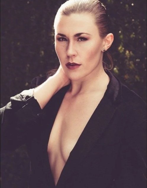 A woman with light skin, blonde hair slicked back, wearing dark lipstick and a black top with an open neckline, standing outdoors with a blurred natural background.