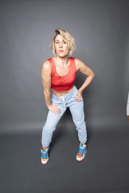 A woman standing against a gray background, wearing a red sleeveless top, light blue sweatpants, and colorful sneakers.