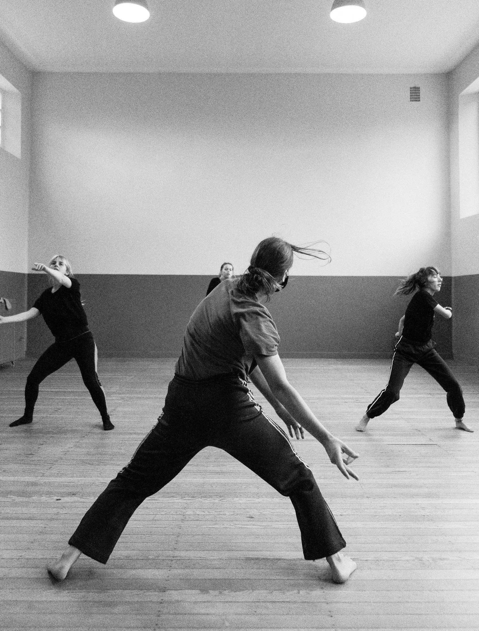 People participating in a yoga or meditation class, with one person in red and others in black, stretching with arms raised, inside a bright room with a large arched window.