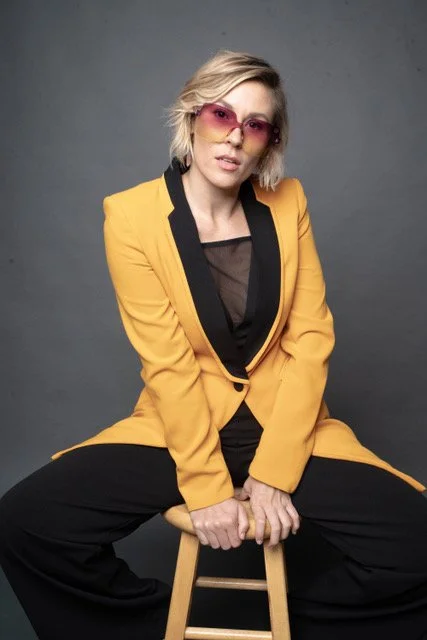 A woman with short blonde hair wearing pink sunglasses and a yellow blazer sitting on a wooden stool against a gray background.