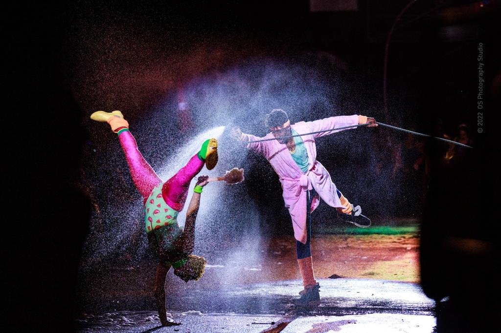 A performer in colorful clothing executes a handstand on stage while a clown in a pink jacket and mask holds a stick and sprays water or glitter towards the performer, creating a dynamic scene with a cloud of spray in the air.