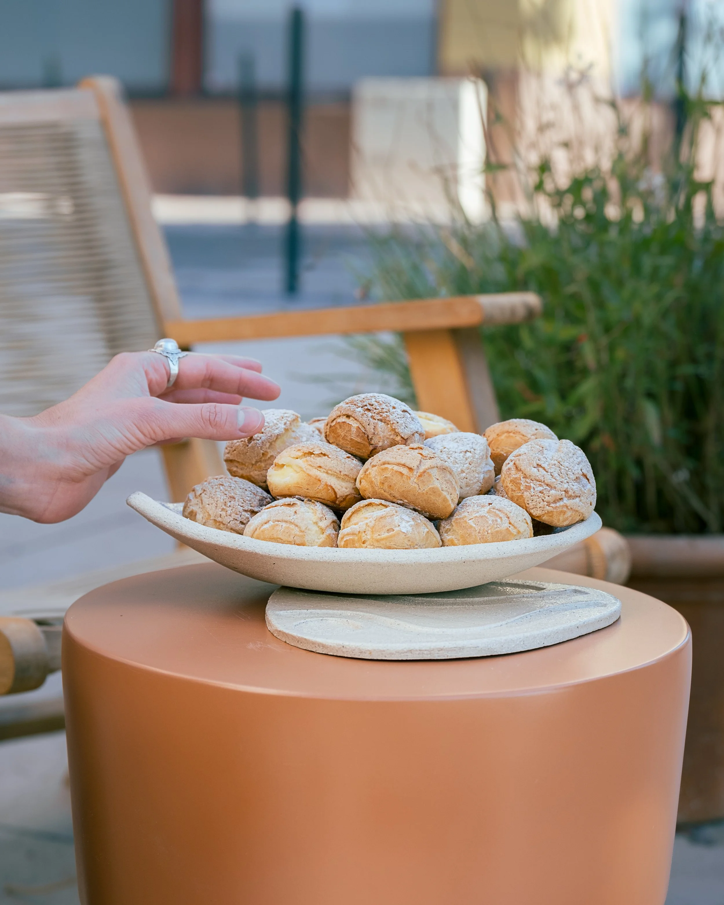 Une main touche une assiette de petits choux ou profiteroles sur une table en extérieur, avec un arrière-plan de mobilier et de végétation.