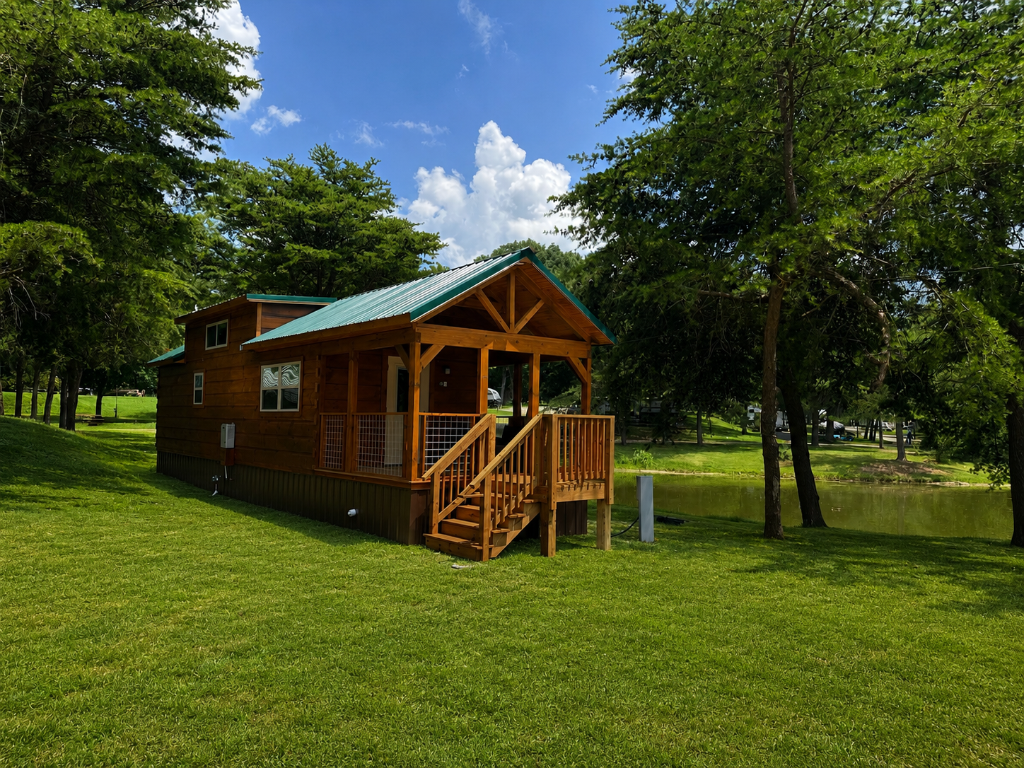 A small wooden cabin with a green metal roof, nestled among green trees by a pond, under a partly cloudy sky.