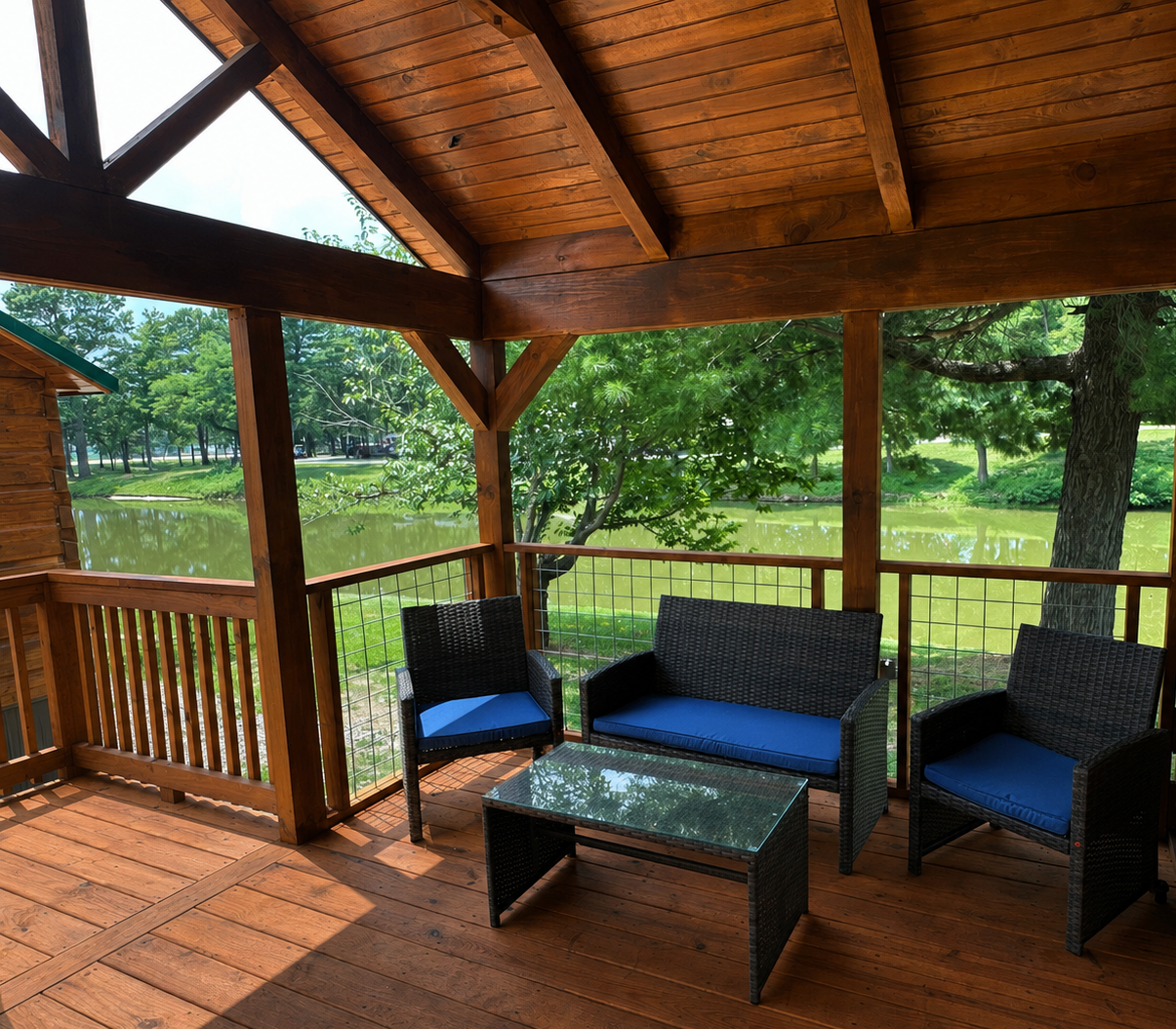 View of a wooden porch overlooking a pond, with black outdoor furniture including two chairs and a small table with blue cushions, surrounded by green trees.