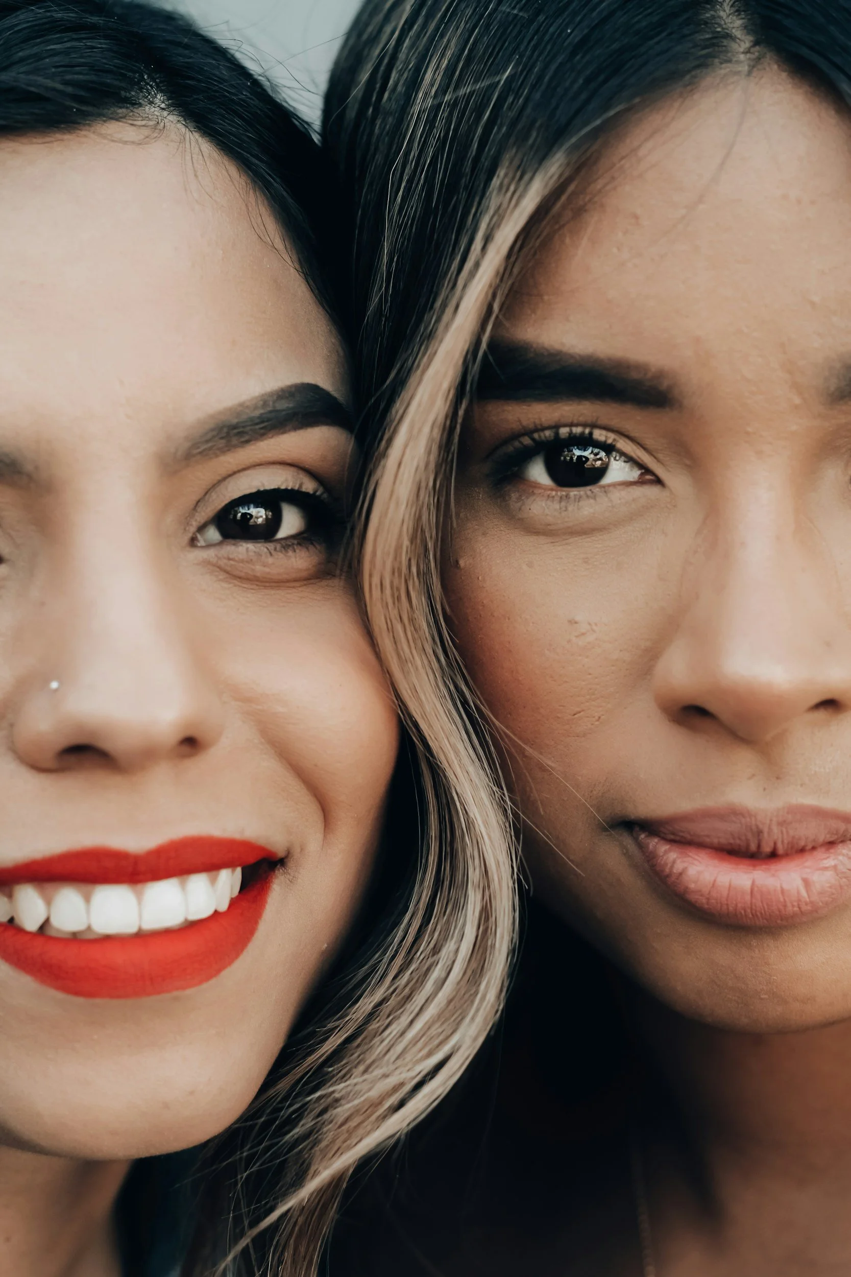 Close-up of two women smiling, showing their faces in a tight frame, with one woman having light skin and red lipstick, and the other having darker skin with natural makeup and a slight smile.