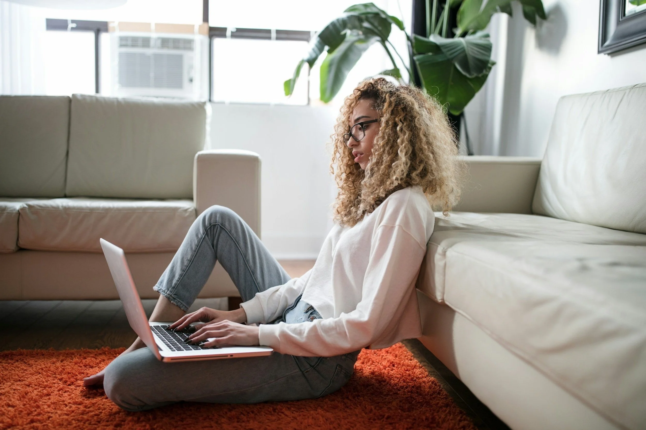 Young woman with curly hair and glasses sitting on the floor leaning against a couch, working on a laptop, in a bright living room with large windows, a large green plant, and an orange rug.