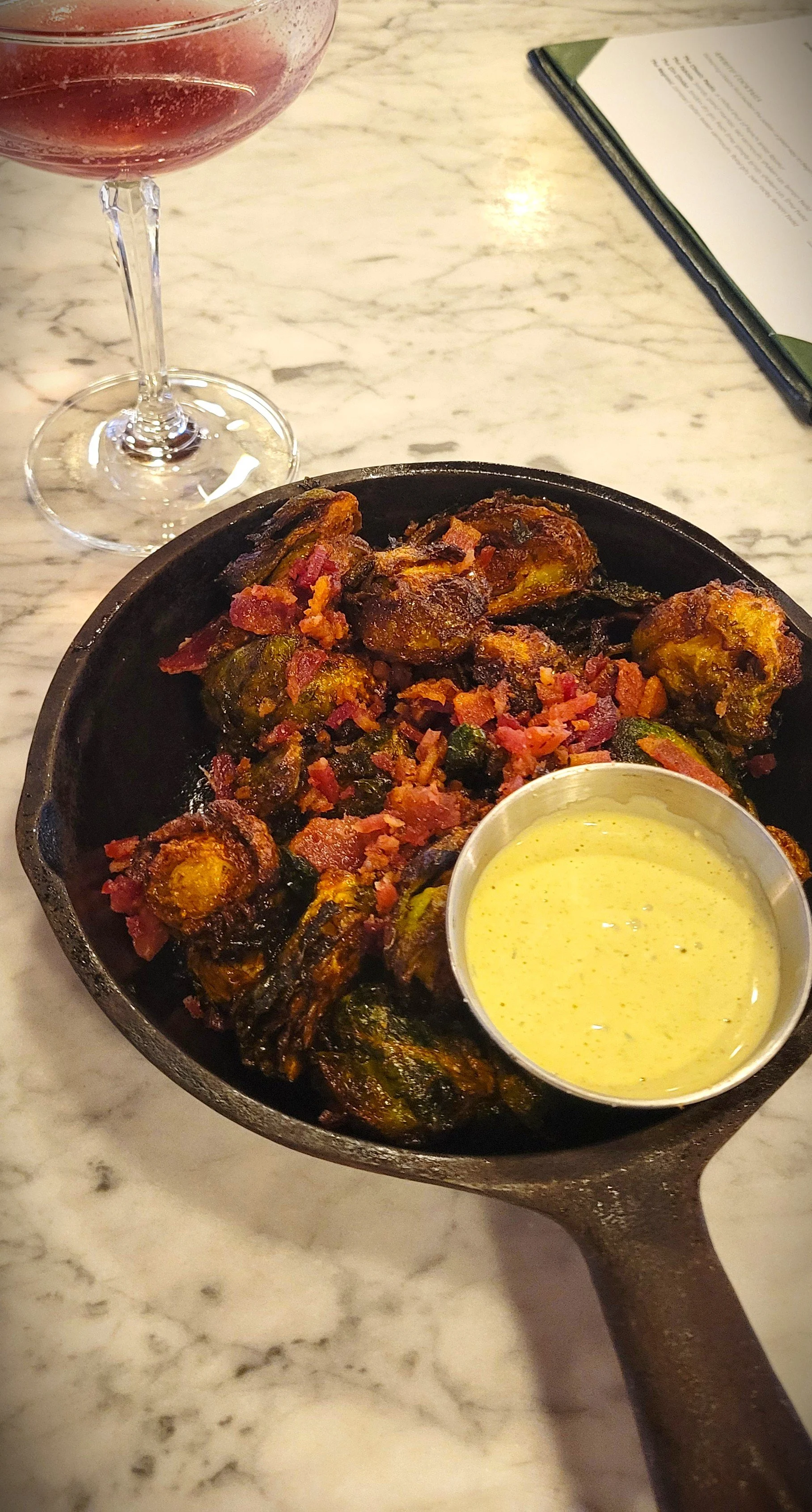 Cooked Brussels sprouts with crispy bacon bits served in a cast iron skillet with a side of yellow mustard sauce, a glass of red wine, and a menu on a marble table.