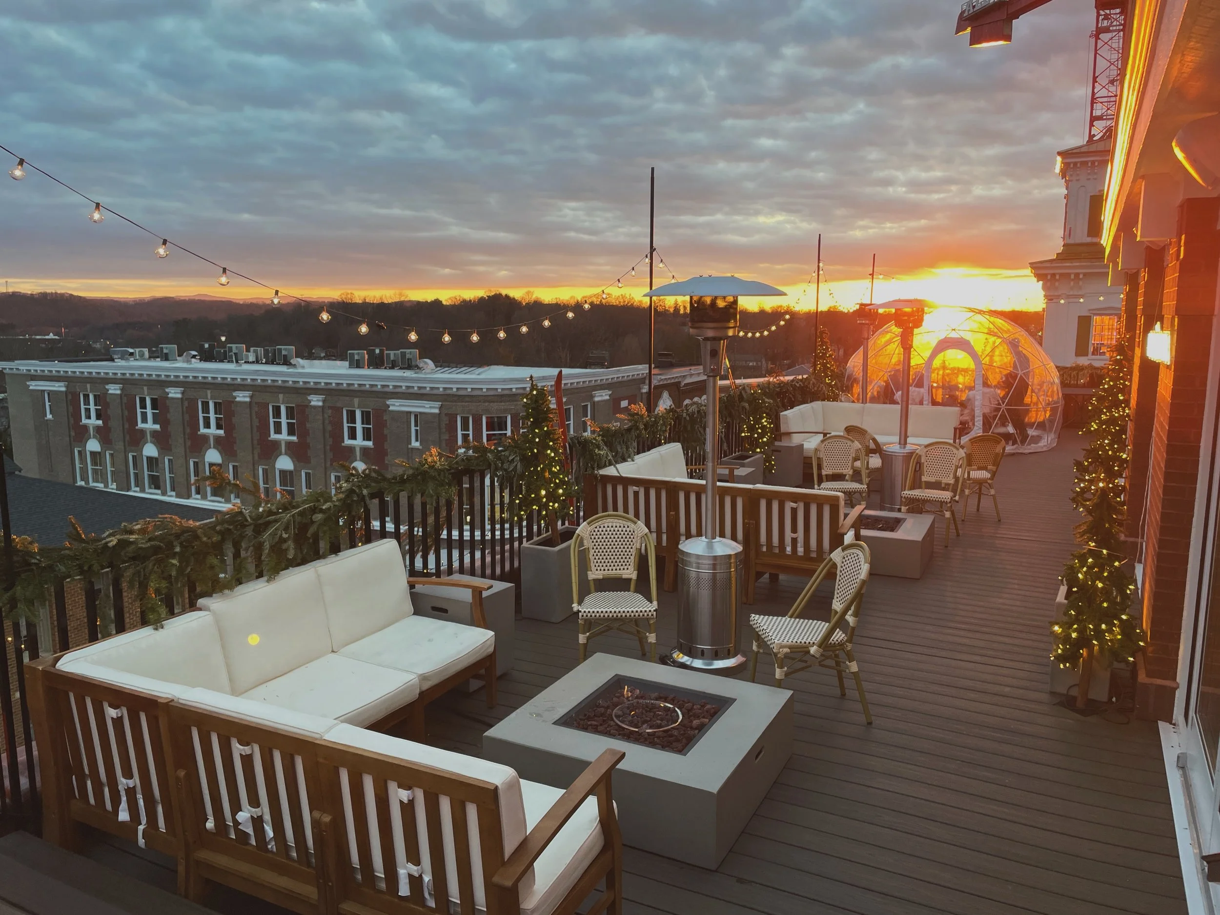Outdoor rooftop patio decorated with string lights and small Christmas trees, with a sunset sky in the background. There are chairs, sofas, patio heaters, and a clear dome structure.